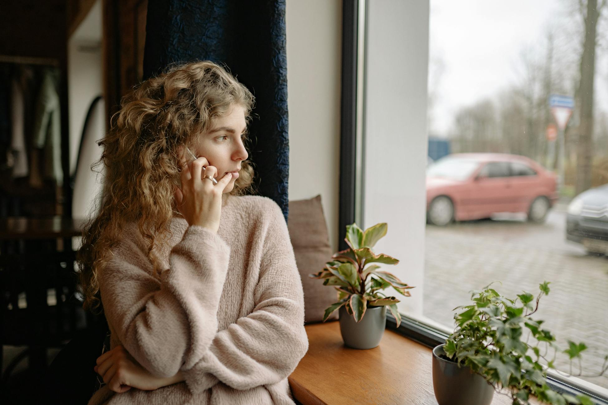 Woman in cozy sweater talking on a phone by the window, surrounded by houseplants, observing outdoors.