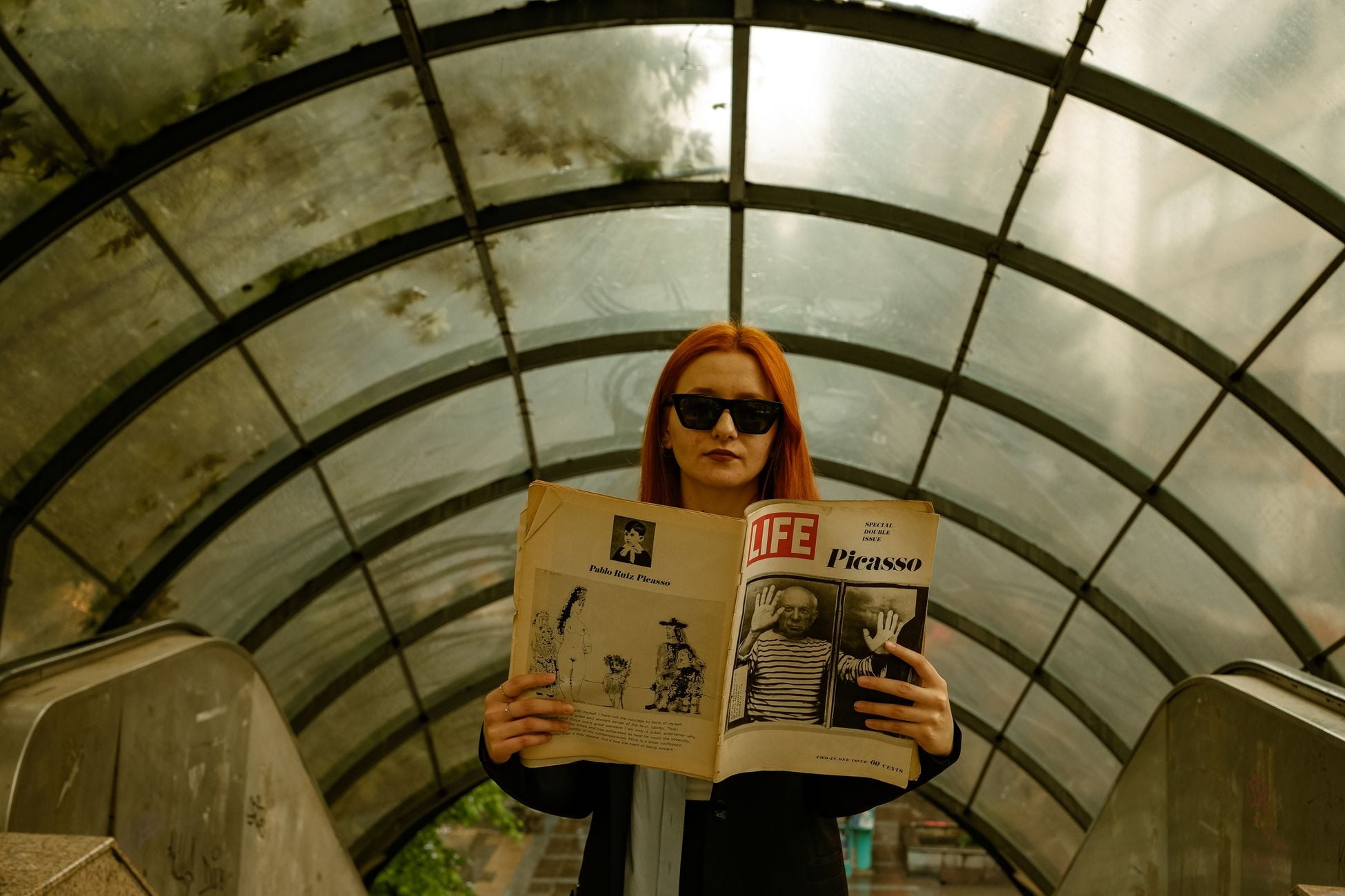 A stylish woman with red hair and sunglasses stands in a glass-ceiling tunnel, holding a vintage magazine.