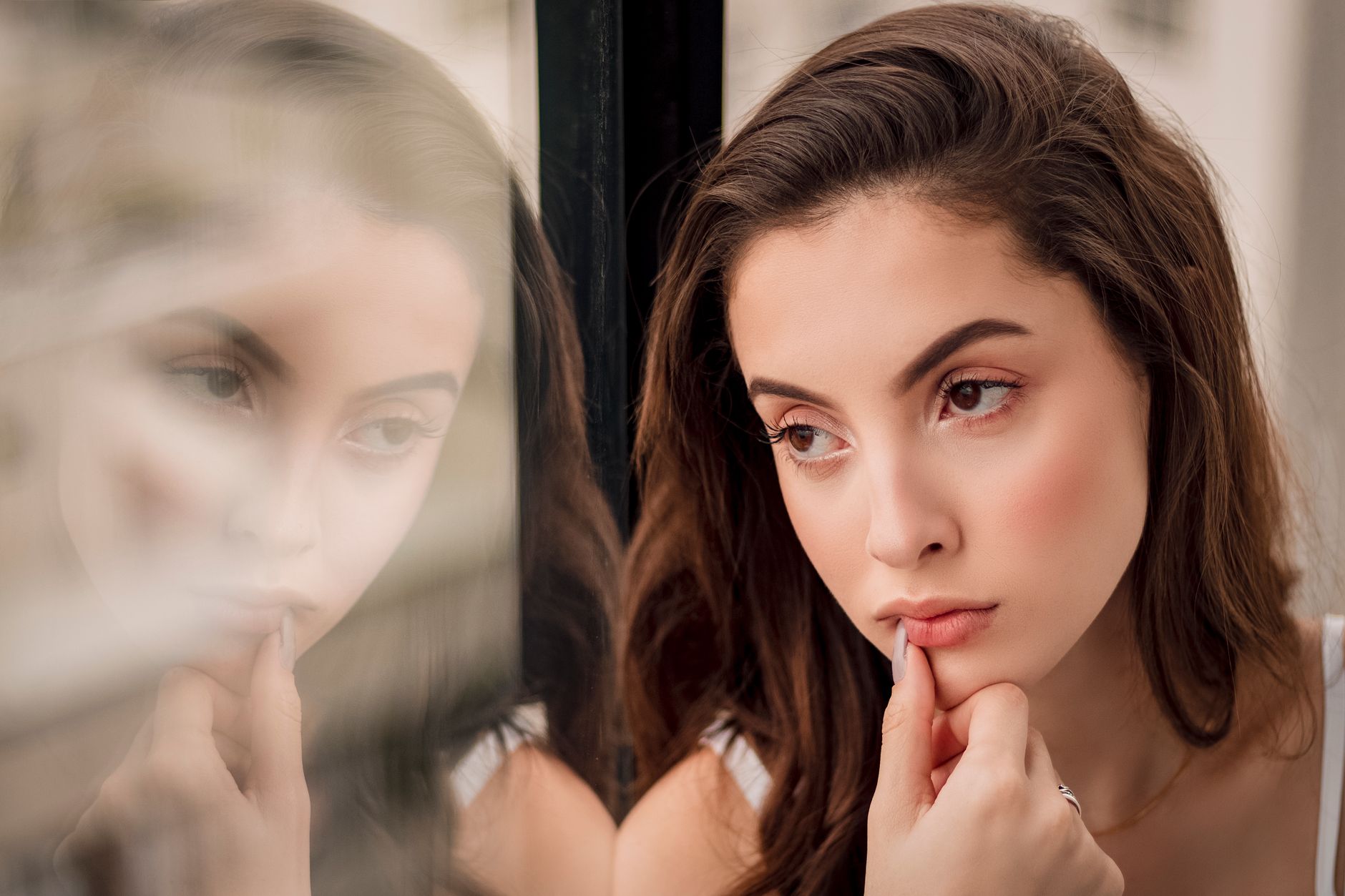 A thoughtful woman beside a window with her face reflected in the glass