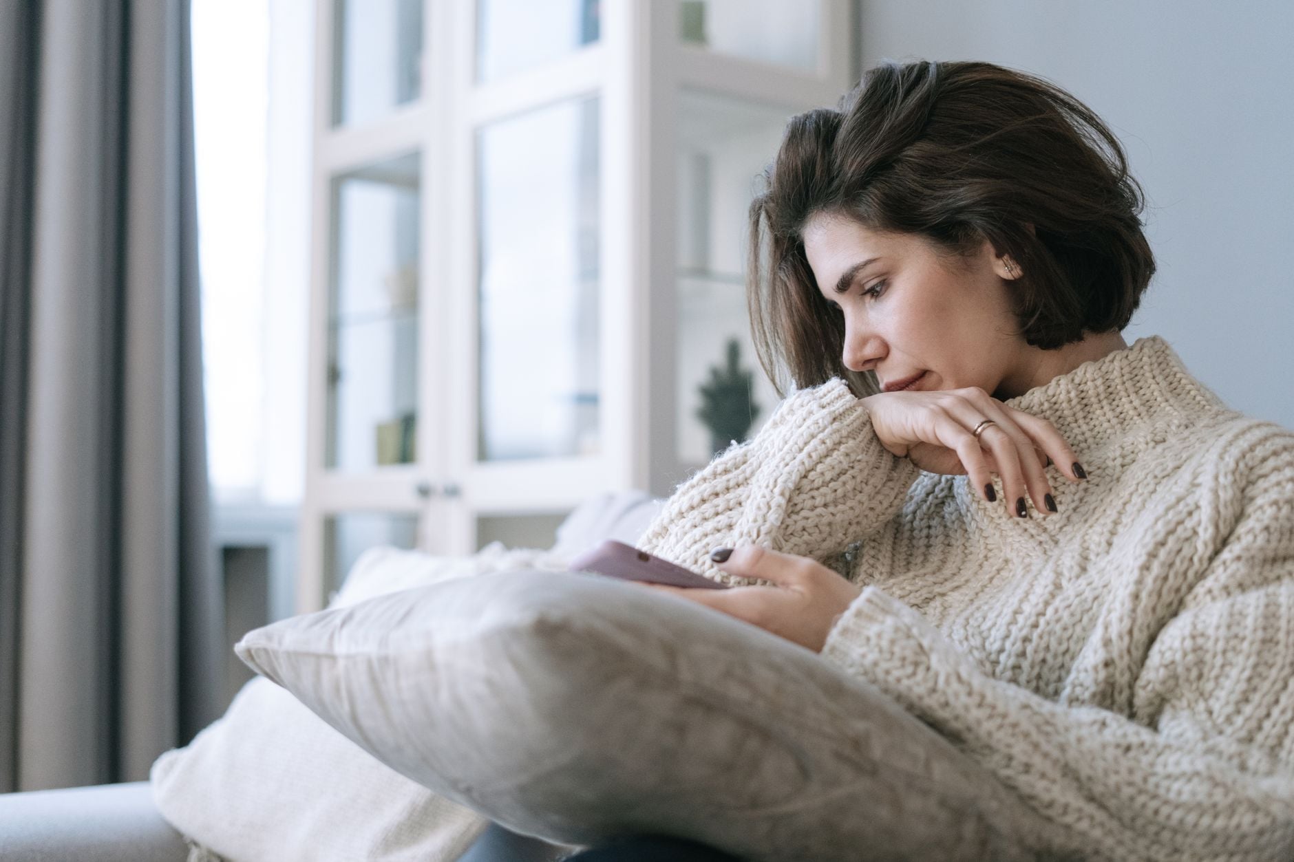 An adult sitting pensively on a couch while using a smartphone at home