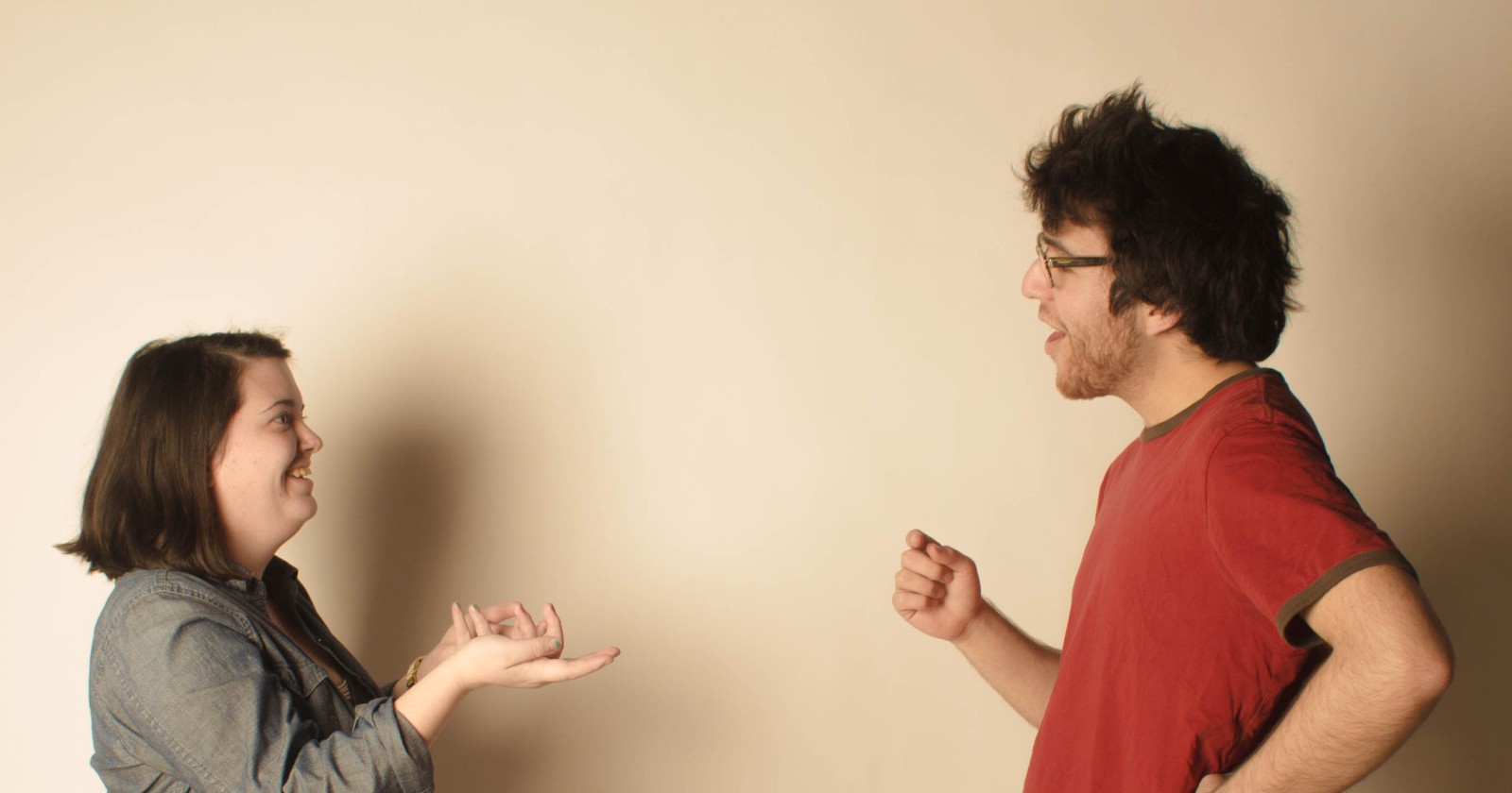 A woman and a man face each other mid-conversation against a plain backdrop, she with open hands, he speaking animatedly