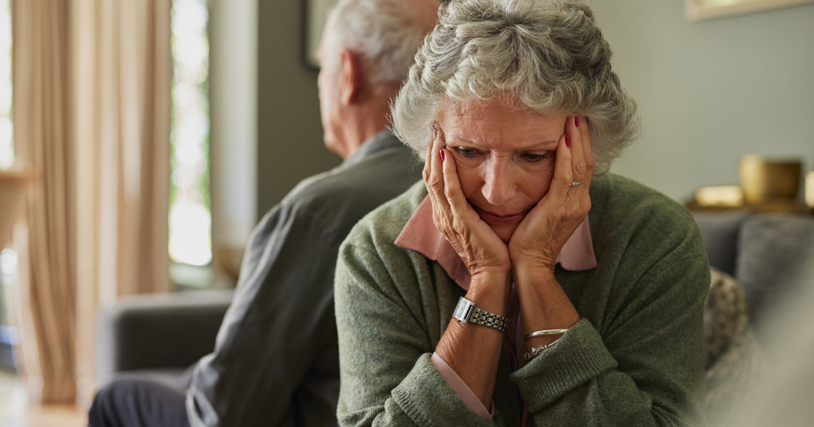 A distressed older woman holding her face while sitting back-to-back with an older man