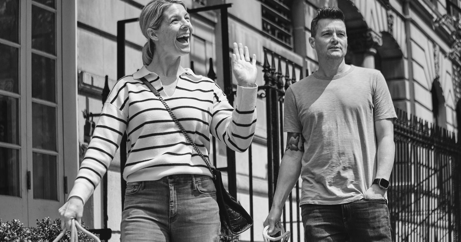 A man and a woman walk together along a city street in black and white. She is laughing with one hand raised; he walks beside her looking ahead.