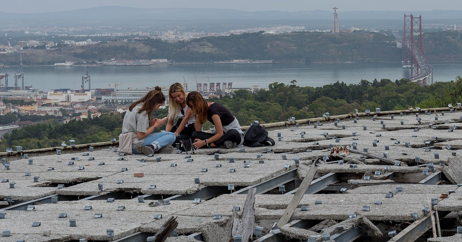 Three young women sit close together on the flat concrete roof of a derelict building, with a river, a long suspension bridge and a hillside city behind them.