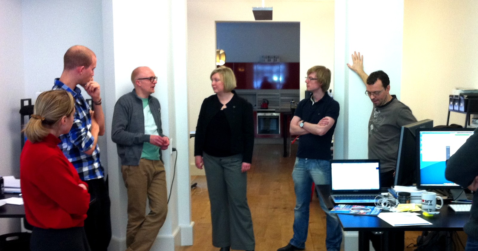 Six adults standing in a small office around a desk with two open laptops during a daily standup meeting; one person stands slightly apart from the cluster.