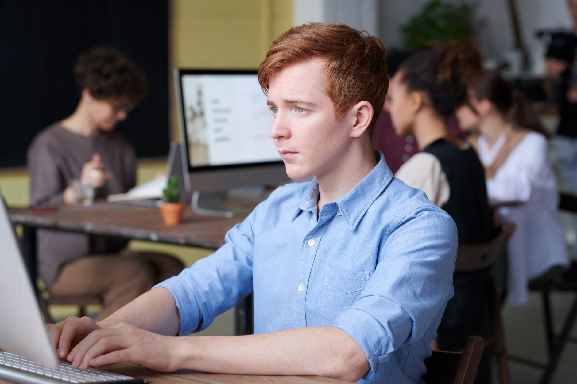 Young professional working at a computer in a busy office with colleagues behind him