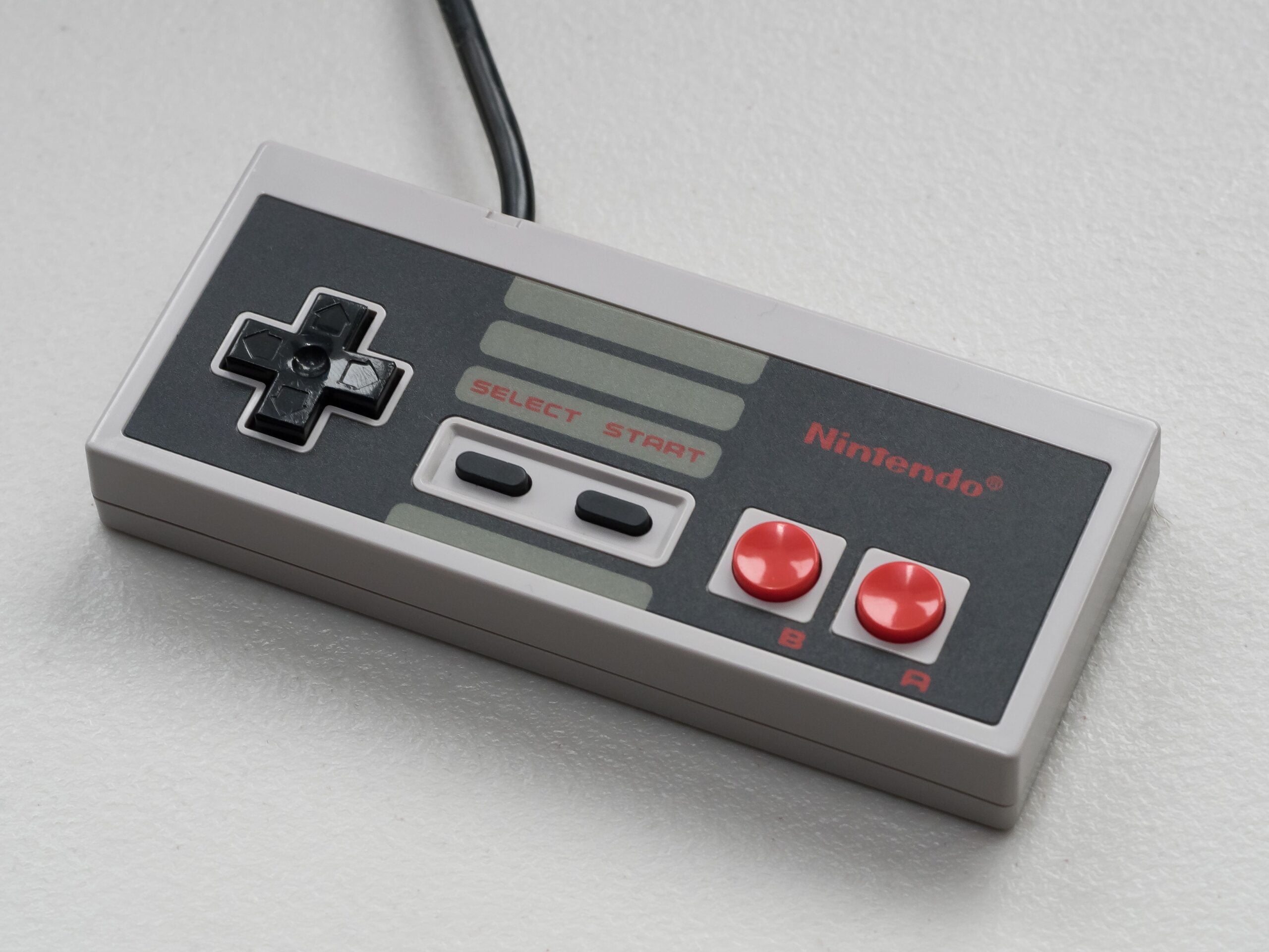 A grey rectangular game controller with a black directional pad and red buttons on a white background