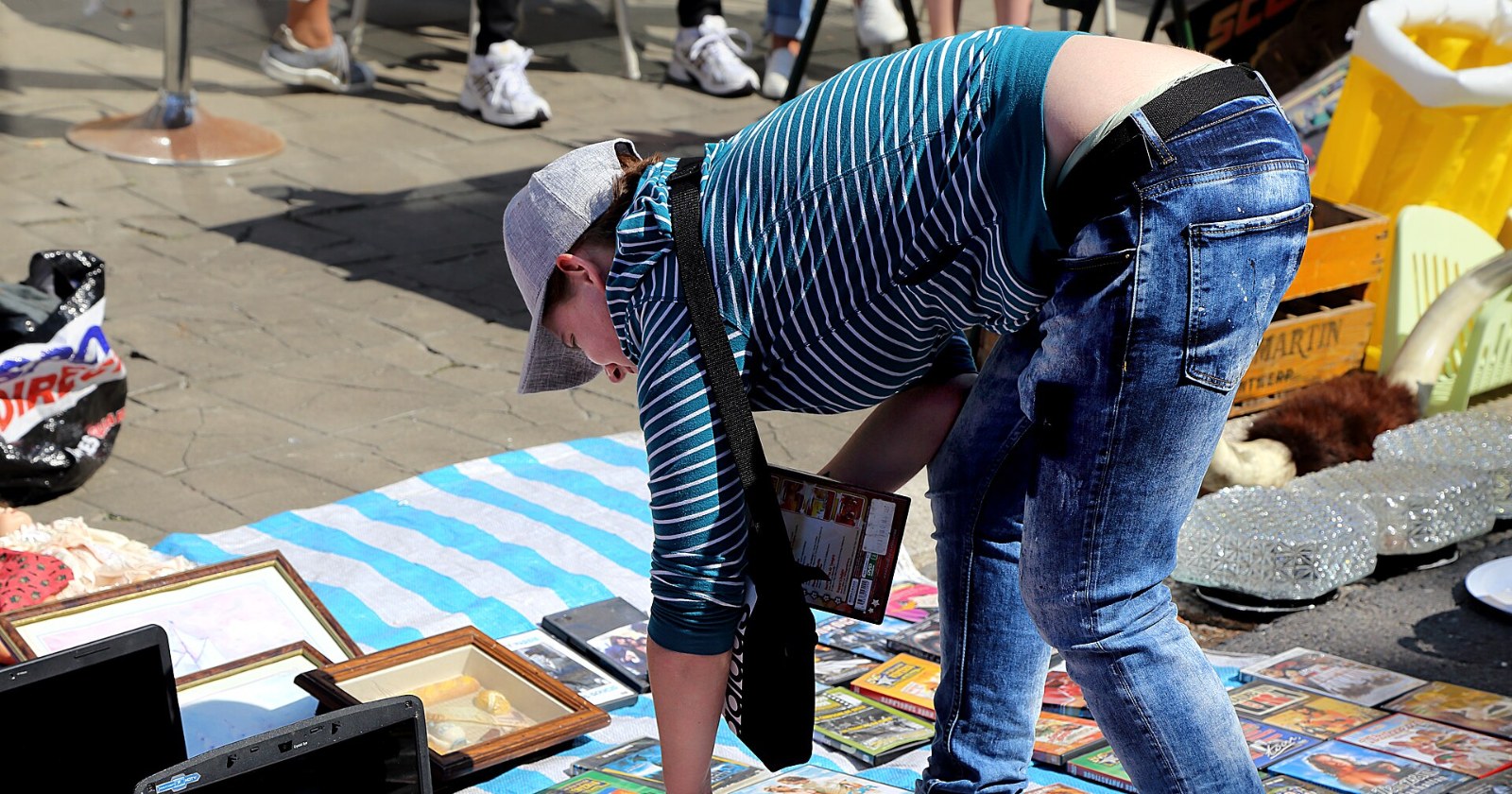 A shopper in a striped top and cap bends over a blanket of secondhand goods, with used laptops laid out in the foreground, at a street flea market