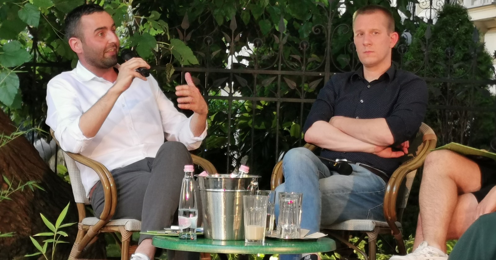 A seated man speaks into a microphone with an open-handed gesture while a second seated man listens with his arms folded, in a garden setting.